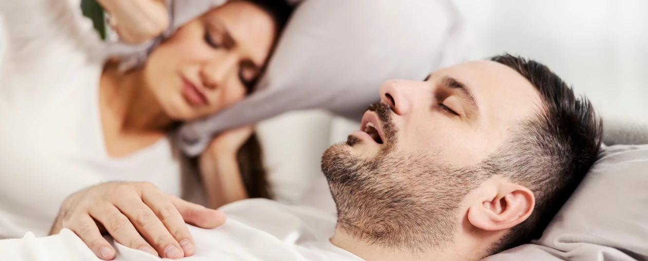 Snoring man in bed with partner covering ears trying to sleep